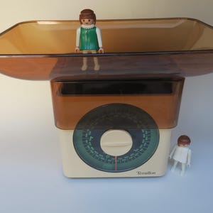 Könnte beinhalten: Eine cremefarbene Vintage-Küchenwaage von Terraillon mit einer großen, transparenten braunen Kunststoffschale. Die Waage verfügt über ein kreisförmiges grünes Zifferblatt mit schwarzen Markierungen und einen zentralen Einstellknopf. Zwei kleine Figuren.
