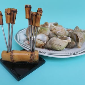 Puede incluir: Un juego de pinchos de bambú con mangos de madera, exhibidos en un soporte negro. Los pinchos están dispuestos en forma de abanico y están listos para ser utilizados para servir comida. Un plato de mariscos cocidos está en el fondo.