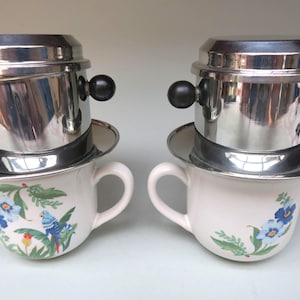 Pode incluir: Dois filtros de café vietnamitas, cada um com um corpo de metal brilhante e alças pretas, assentam sobre canecas de cerâmica brancas. As canecas são decoradas com desenhos florais e de pássaros azuis. Método tradicional.