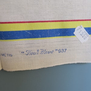 Puede incluir: Una toalla de cocina de lino blanco con rayas rojas, amarillas y azules. La toalla tiene el texto "Toile M&eacute;tis" y "Fleur Bleue" 937 impreso en ella. Una etiqueta de precio blanca con el texto "F13.00" y "11020 68" est&aacute; unida a la toalla.