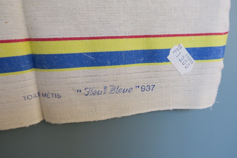 Puede incluir: Una toalla de cocina de lino blanco con rayas rojas, amarillas y azules. La toalla tiene el texto "Toile M&eacute;tis" y "Fleur Bleue" 937 impreso en ella. Una etiqueta de precio blanca con el texto "F13.00" y "11020 68" est&aacute; unida a la toalla.