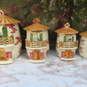 Könnte beinhalten: Ein Set aus vier keramikförmigen Behältern in Form eines Hauses. Jedes hat ein gelbes Dach, einen goldfarbenen Balkon und rote Ziegelakzente. Die Behälter sind cremefarben mit grünen Fensterläden und einer kleinen Tür. Floral Hintergrund.