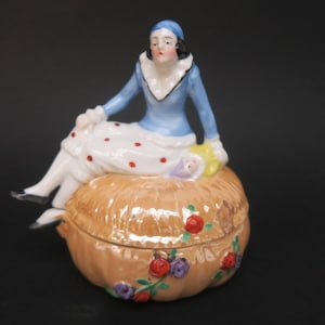 Könnte beinhalten: Eine Porzellan-Schmuckdose mit einer sitzenden Frauenfigur darauf. Die Frau trägt ein blaues und weißes Kleid mit roten Punkten. Die Dose ist mit Blumen verziert und hat eine goldfarbene Oberfläche.