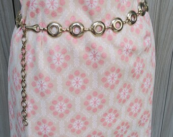 May include: A gold chain belt with oval-shaped links. The belt is worn on a dress with a pink and white floral pattern.
