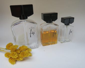 May include: Three clear glass perfume bottles with black square caps. The bottles are labeled "Fiji" in a script font. One bottle is filled with amber liquid. A bundle of yellow dried flowers is in the foreground.