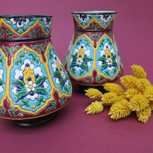 May include: Two decorative ceramic vases with intricate floral patterns in shades of blue, yellow, green, and red on a burgundy background. The vases are sitting on a burgundy surface with a bouquet of dried yellow flowers.