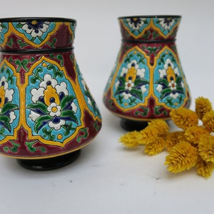 May include: Two small, decorative ceramic vases with a colorful floral pattern. The vases are a deep red color with blue, green, yellow, and white accents. The vases are sitting on a white surface.