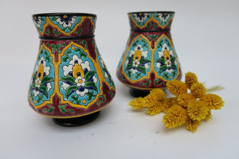 May include: Two small, decorative ceramic vases with a colorful floral pattern. The vases are a deep red color with blue, green, yellow, and white accents. The vases are sitting on a white surface.