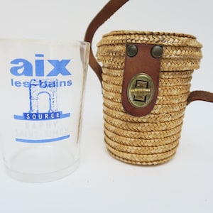May include: A clear glass with blue text that reads "aix les bains" and a straw woven bag with a brown leather strap and clasp. The bag is cylindrical and has a brown leather strap.