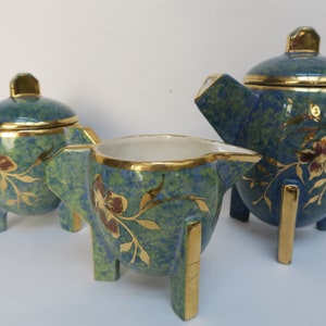 Op de afbeelding: Een driedelige keramische theeset met een blauwgroene gevlekte afwerking, geaccentueerd met gouden randen en bloemmotieven. De set bevat een theepot, een suikerpot en een melkkan, elk met een unieke vorm en drie kleine pootjes.