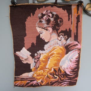 Peut inclure: Tapisserie au point de croix représentant une femme lisant un livre. L'œuvre présente une femme en robe jaune, assise dans une chaise, avec un fond marron et rose. La tapisserie a une bordure marron et est probablement vintage.