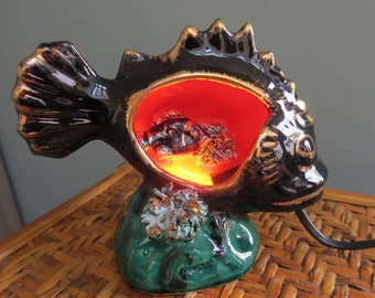 Può includere: Una lampada a forma di pesce in ceramica nera con interno rosso e base verde. Il pesce ha dettagli dorati e una luce all'interno.