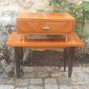 Puede incluir: Una mesita de noche de estilo moderno de mediados de siglo con un cajón. La mesita está hecha de madera con un acabado cálido de color marrón rojizo. El cajón tiene un patrón de espiga y un tirador plateado. Las patas son negras con puntas doradas. La mesita mide aproximadamente 61 cm de alto.