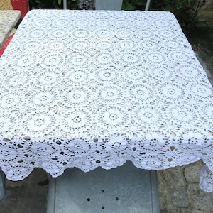 Peut inclure: Une nappe carrée blanche présentant un motif complexe au crochet composé de cercles répétitifs et de détails en dentelle ajourée. Le tissu a un aspect délicat et texturé avec des bords festonnés qui retombent sur les côtés de la table.