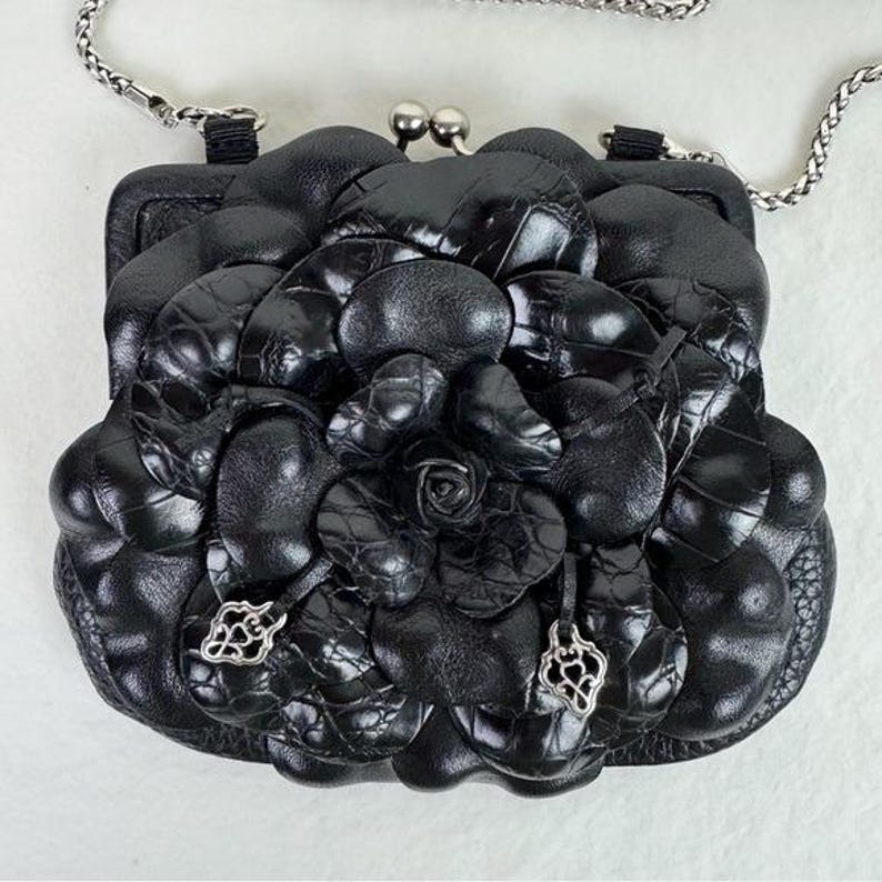 Może przedstawiać: Czarna sk&oacute;rzana torebka z kwiatowym wzorem. Torebka posiada srebrny łańcuszek i zapięcie na zatrzask. Kwiatowy wz&oacute;r wykonany jest ze sk&oacute;ry o fakturze, z r&oacute;żą pośrodku i dekoracyjnymi srebrnymi zawieszkami.