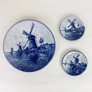 May include: Three blue and white decorative plates depicting a windmill scene. The plates are of varying sizes, with the largest being round and the smaller two also round. The plates feature a detailed illustration of windmills, water, and buildings.