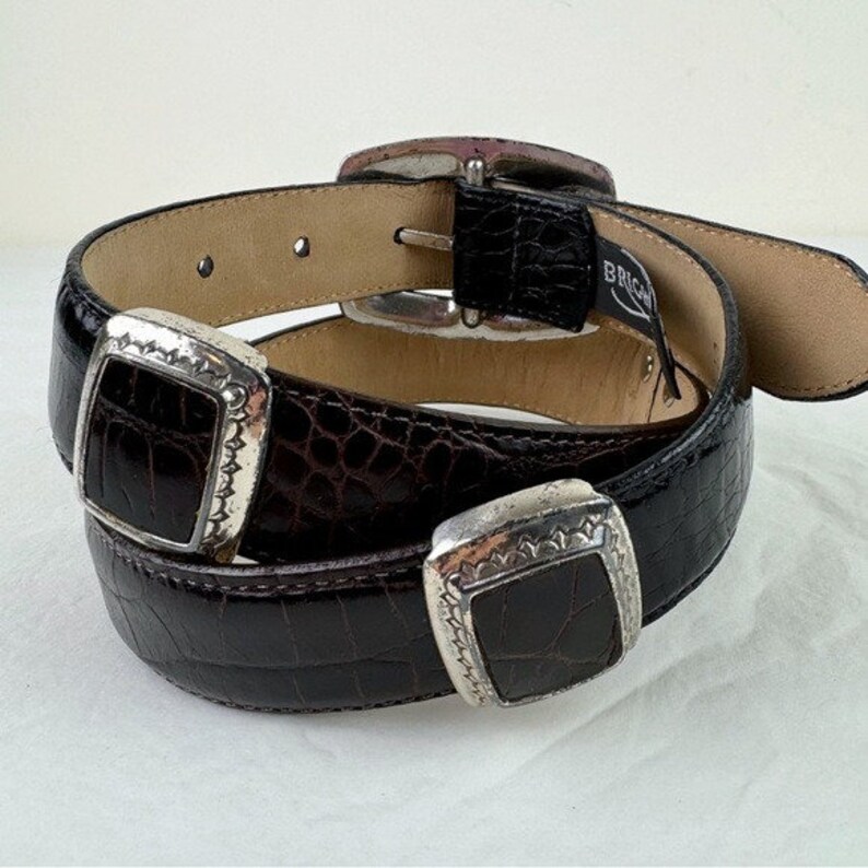 May include: A brown leather belt with two silver buckles. The belt is made of crocodile embossed leather.