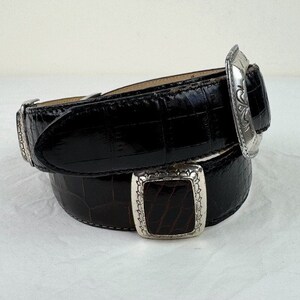 May include: A black leather belt with a silver buckle. The buckle has a detailed design and is a statement piece.