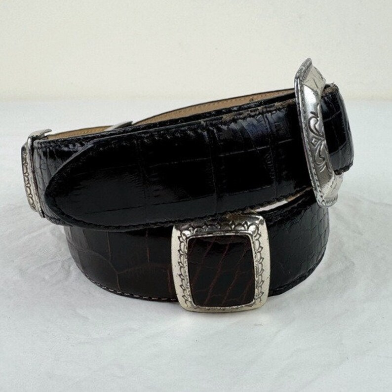 May include: A black leather belt with a silver buckle. The buckle has a detailed design and is a statement piece.