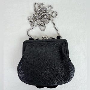 Może przedstawiać: Czarna sk&oacute;rzana portmonetka ze srebrnym łańcuszkiem. Portmonetka ma teksturowaną powierzchnię i zapięcie na zatrzask. Łańcuszek ma delikatny, tkany wz&oacute;r. Portmonetka jest mała i kompaktowa.