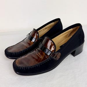 May include: A pair of brown and black leather loafers with a black suede trim and a small buckle detail. The shoes have a low heel and a pointed toe.