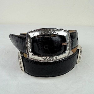 May include: A black leather belt with a silver buckle featuring an ornate design. The belt is folded in half, showing the buckle and the leather.