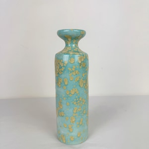 May include: A tall, cylindrical vase with a turquoise glaze and speckled, golden-yellow accents. The vase has a flared top and a narrow neck. The ceramic vessel is a decorative item for the home.