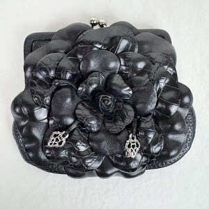 Może przedstawiać: Czarna sk&oacute;rzana portmonetka z kwiatowym wzorem. Portmonetka ma duży, teksturowany kwiat z r&oacute;żą pośrodku. Portmonetka ma srebrne zapięcie i dekoracyjne srebrne zawieszki.