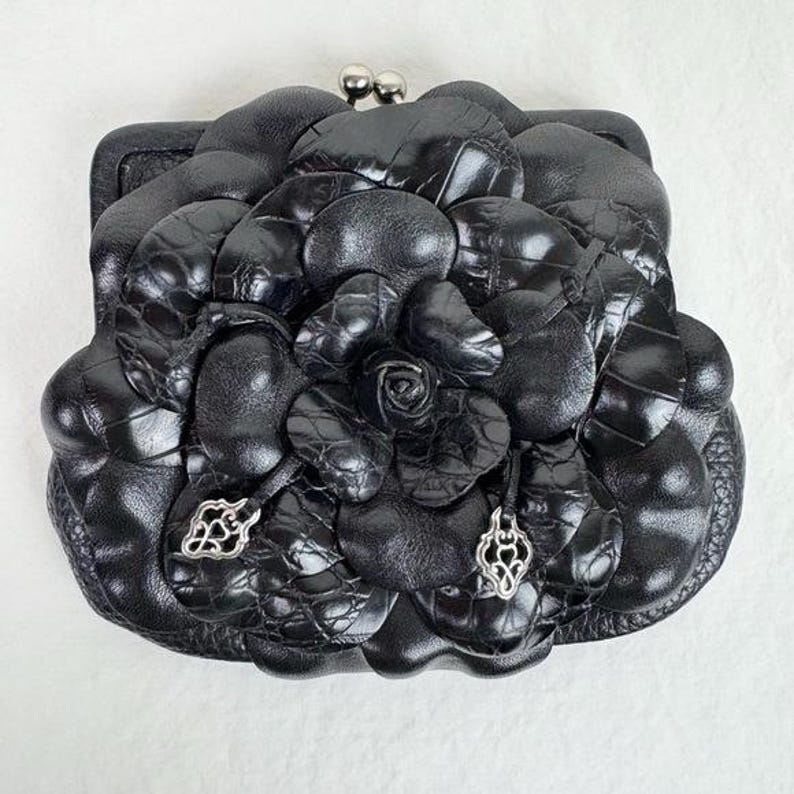 Może przedstawiać: Czarna sk&oacute;rzana portmonetka z kwiatowym wzorem. Portmonetka ma duży, teksturowany kwiat z r&oacute;żą pośrodku. Portmonetka ma srebrne zapięcie i dekoracyjne srebrne zawieszki.