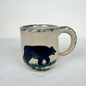 May include: A beige ceramic mug featuring a black bear illustration and green grass detail. The rim and handle have a green trim. The mug is a standard size for tea or coffee.
