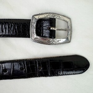 May include: A black leather belt with a silver buckle featuring an ornate design. The belt has a crocodile-embossed pattern.