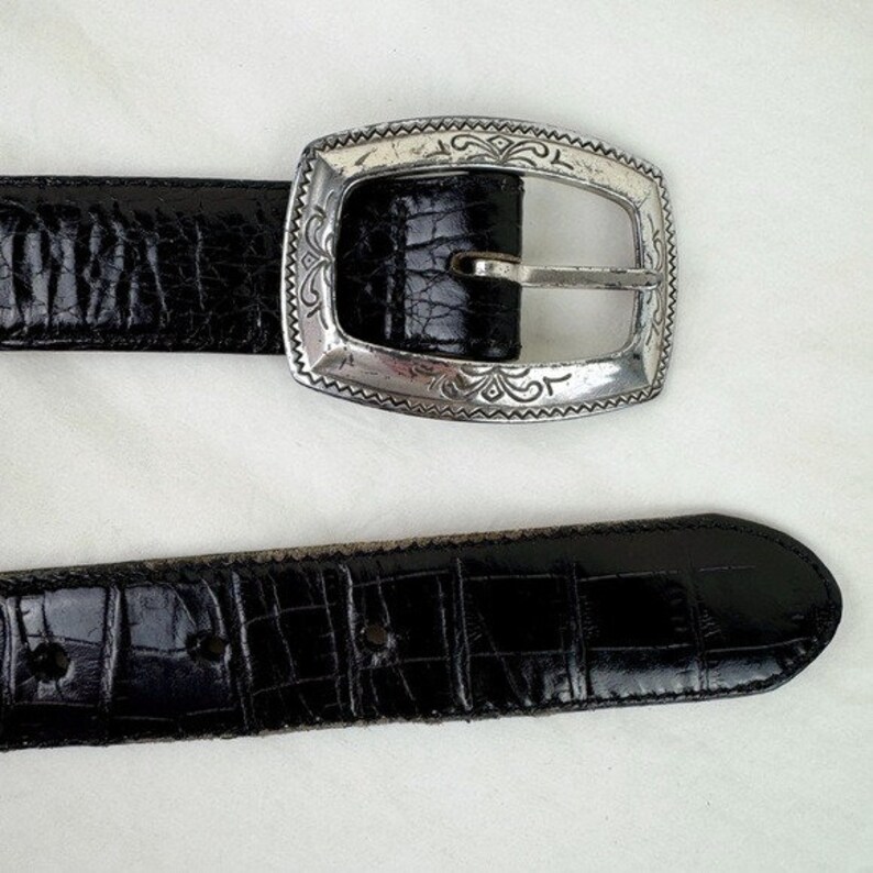 May include: A black leather belt with a silver buckle featuring an ornate design. The belt has a crocodile-embossed pattern.