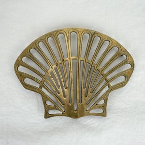 Puede incluir: Un salvamanteles en forma de abanico de color latón, con un diseño calado. El salvamanteles presenta un patrón de concha con líneas curvas que irradian desde el centro. El metal tiene un aspecto ligeramente envejecido.