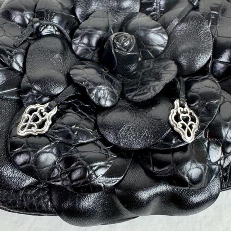 Może przedstawiać: Zbliżenie czarnej sk&oacute;rzanej torebki z kwiatowym wzorem. Torba ma teksturowany, wytłoczony wz&oacute;r i jest ozdobiona srebrnymi zawieszkami. Projekt zawiera r&oacute;żę i kształty przypominające płatki.