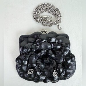 Może przedstawiać: Czarna sk&oacute;rzana kopert&oacute;wka z kwiatowym wzorem i srebrnym łańcuszkiem. Kopert&oacute;wka posiada zapięcie typu kiss-lock i ozdobne srebrne zawieszki. Kwiatowy wz&oacute;r wykonany jest z teksturowanej sk&oacute;ry. Srebrny łańcuszek ma około 122 cm długości.