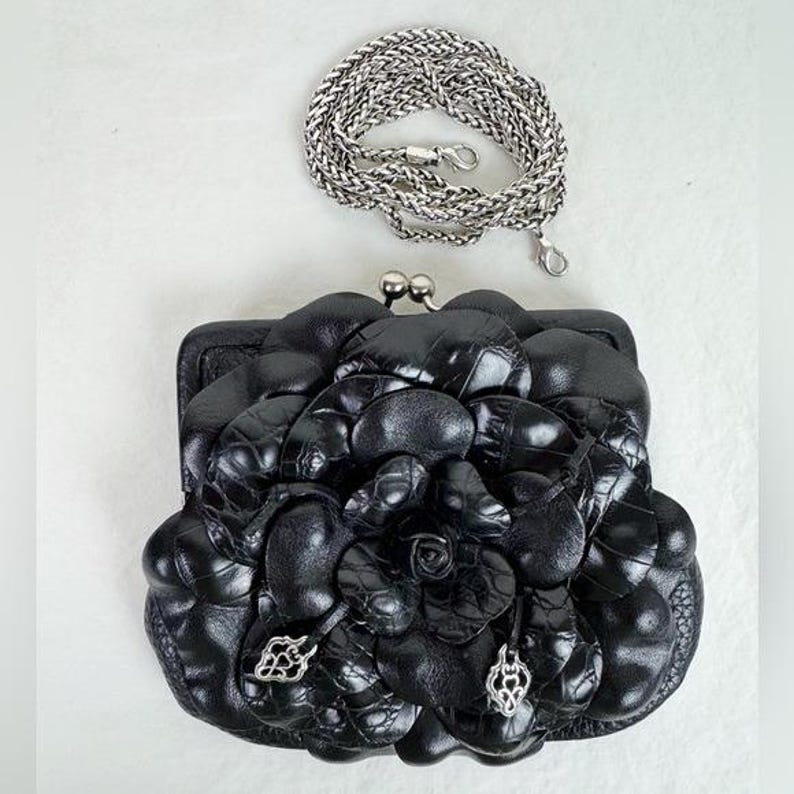 Może przedstawiać: Czarna sk&oacute;rzana kopert&oacute;wka z kwiatowym wzorem i srebrnym łańcuszkiem. Kopert&oacute;wka posiada zapięcie typu kiss-lock i ozdobne srebrne zawieszki. Kwiatowy wz&oacute;r wykonany jest z teksturowanej sk&oacute;ry. Srebrny łańcuszek ma około 122 cm długości.