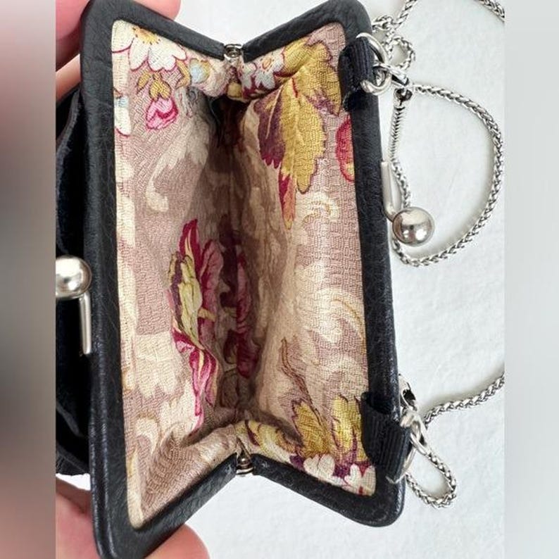 Może przedstawiać: Czarna sk&oacute;rzana portmonetka ze srebrnym łańcuszkiem. Wnętrze ma kwiatowy wz&oacute;r w odcieniach r&oacute;żu, ż&oacute;łci i zieleni na neutralnym tle. Portmonetka ma metalowe zapięcie i mały srebrny detal w kształcie kuli.