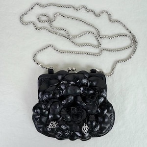 Może przedstawiać: Czarna sk&oacute;rzana kopert&oacute;wka ze srebrnym łańcuszkiem. Kopert&oacute;wka ma kwiatowy wz&oacute;r z przodu i srebrne zapięcie. Z przodu przymocowane są dwa srebrne charmsy.