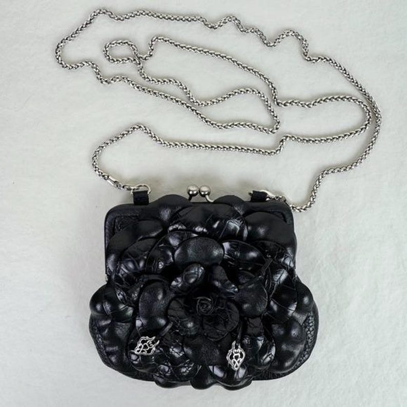 Może przedstawiać: Czarna sk&oacute;rzana kopert&oacute;wka ze srebrnym łańcuszkiem. Kopert&oacute;wka ma kwiatowy wz&oacute;r z przodu i srebrne zapięcie. Z przodu przymocowane są dwa srebrne charmsy.