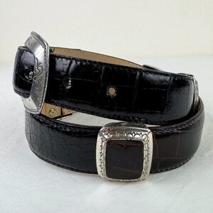 May include: A black alligator leather belt with a silver buckle featuring an ornate design. The buckle has a square shape with a smaller square inset.