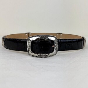 May include: A black leather belt with a silver buckle featuring an ornate design. The belt has a crocodile-embossed texture.