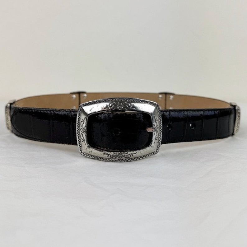 May include: A black leather belt with a silver buckle featuring an ornate design. The belt has a crocodile-embossed texture.