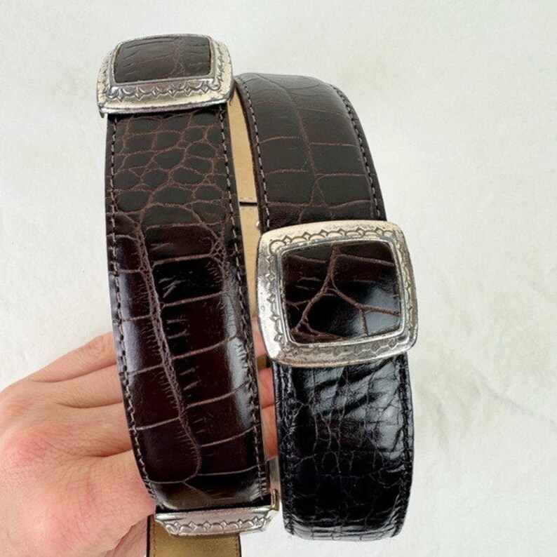 May include: Two brown leather belts with silver buckles. The buckles have a square shape with a decorative border. The belts have a crocodile pattern.
