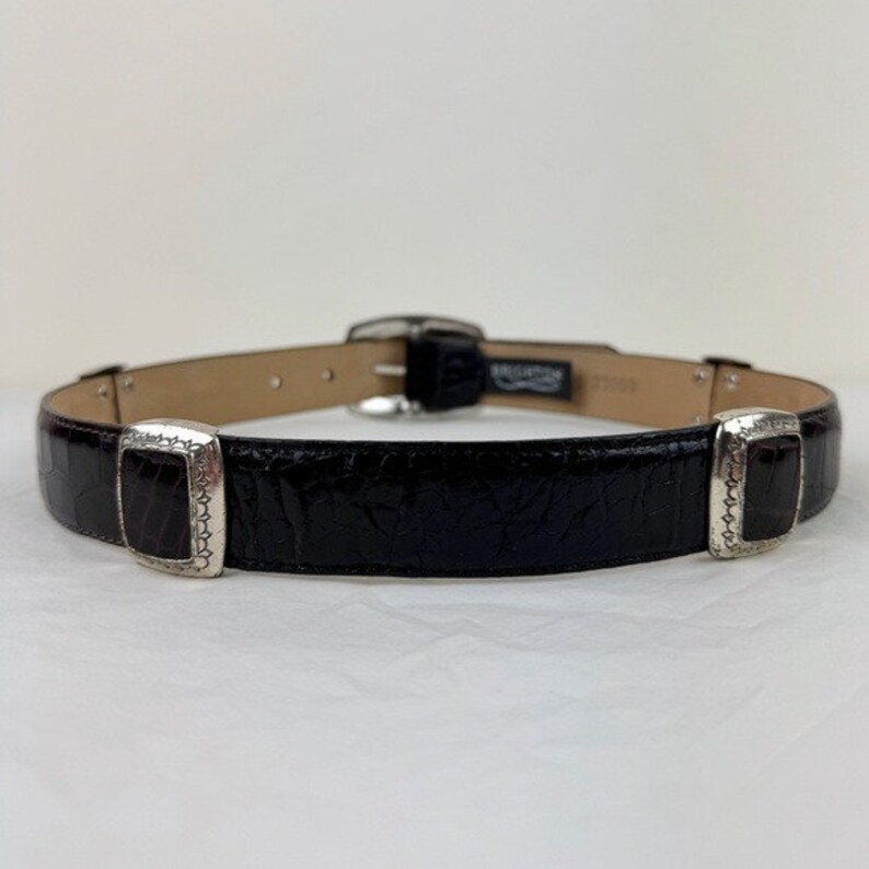 May include: A black leather belt with silver-toned buckles. The belt has a textured, alligator-like pattern.