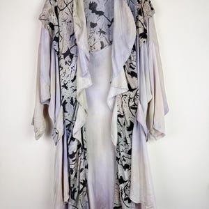 May include: A long, flowing, light purple and white silk robe with black abstract designs. The robe has a shawl collar and is open in the front. The label "Rebel to Plaid" is visible on the collar.