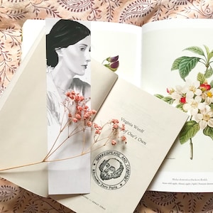 Könnte beinhalten: Eine Nahaufnahme eines Buches mit dem Titel "A Room of One's Own" von Virginia Woolf. Das Buch ist geöffnet und hat ein Lesezeichen mit einer Schwarzweißillustration einer Frau. Das Buch liegt auf einer gemusterten Stoffoberfläche.