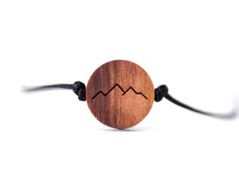 May include: A wooden bracelet with a black cord and a round wooden charm featuring a mountain range design.