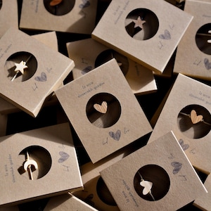 May include: A collection of small, brown cardboard boxes with a heart cutout on the front. Each box contains a wooden charm bracelet with a different design, including a heart, star, and crescent moon. The boxes are labeled with the text "Iz srca slovenskih gozdov", which translates to "From the heart of Slovenian forests".