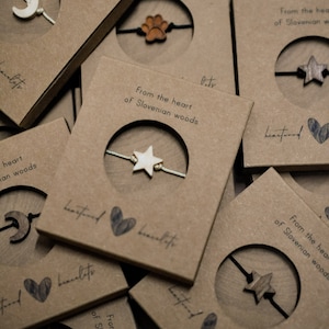Può includere: Una collezione di braccialetti con ciondoli in legno esposti su confezioni di cartone marrone. I braccialetti presentano varie forme, tra cui una stella, una luna e un'impronta di zampa. La confezione include il testo "From the heart of Slovenian woods."