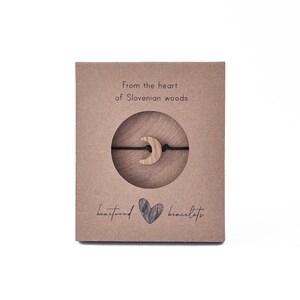 May include: A brown cardboard box with a black string bracelet with a wooden crescent moon charm. The box has the text "From the heart of Slovenian woods" and "heartwood bracelets".